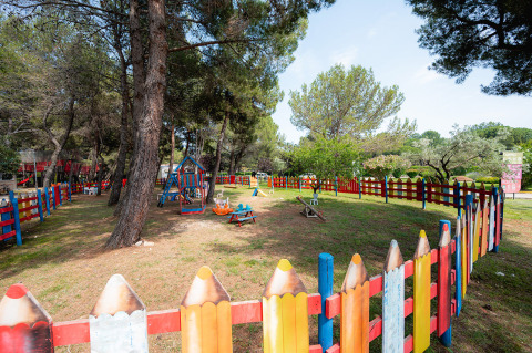 Colorful fenced playground area in a wooded park at Bi Village holiday park, Istria, Croatia, on a sunny day.