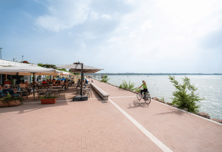 A cyclist rides along a lakeside promenade near an outdoor café with people dining in Veneto, Italy.