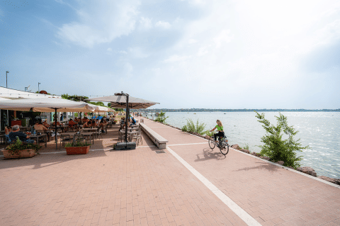 Un ciclista pasea junto al lago cerca de una cafetería al aire libre, con gente comiendo en Veneto, Italia.