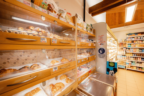 Bakery section of a store with croissants and pastries in wooden shelves, dairy products in the background.