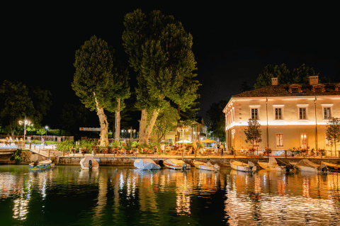 Vue nocturne avec bateaux amarrés et bâtiments illuminés au bord de l’eau à Campeggio del Garda, Vénétie, Italie.
