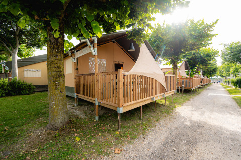Safari tent Villatent Outback met houten veranda op Campeggio del Garda in Italië, onder schaduwrijke bomen.