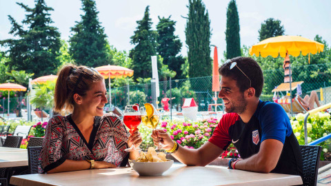 Una pareja brinda con bebidas en una terraza de Campeggio Gasparina, parque vacacional en Véneto, Italia.