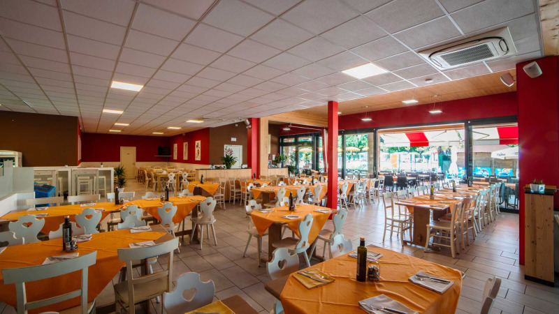 Indoor restaurant seating at Campeggio Gasparina, Veneto, Italy, with orange tablecloths and large windows.