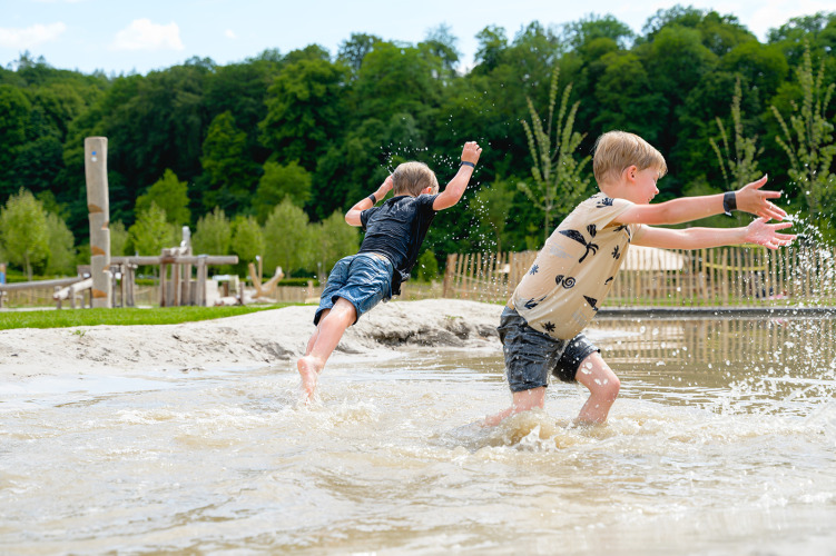 Zwei Jungen spielen und planschen im Wasser eines Campingplatzes, umgeben von Bäumen in Limburg, Niederlande.