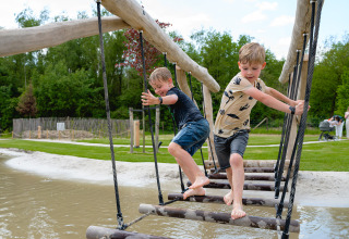 Dos niños juegan en un puente de cuerda sobre agua en Camping 't Geuldal en Limburg, Países Bajos.