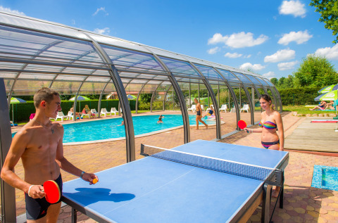 Dos jóvenes juegan al tenis de mesa junto a una piscina cubierta en Camping Au Clos de La Chaume, Francia.