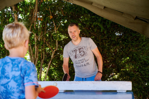 Un hombre y un niño juegan al ping-pong al aire libre bajo una lona en un camping de Grand Est, Francia.