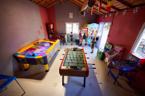 Bambini che giocano in una sala giochi colorata con calcio balilla, air hockey e cabinati in un villaggio vacanze francese.