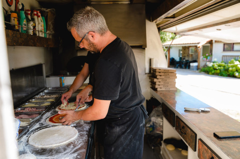 Due persone preparano pizze in cucina con ingredienti, scatole per pizza e area esterna sullo sfondo.