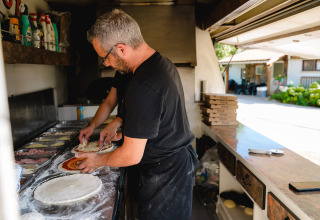 Due persone preparano pizze in cucina con ingredienti, scatole per pizza e area esterna sullo sfondo.