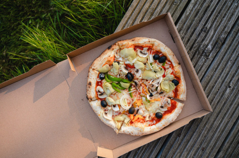 Pizza mit Gemüse wie Oliven, Artischocken und Pilzen in einer Schachtel, draußen auf Holzterrasse und Gras.
