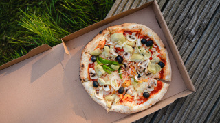 Pizza vegetariana con aceitunas, alcachofas y champiñones en caja sobre tarima de madera junto al césped.