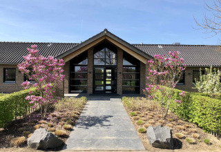 Entrance of Camping Betuwe showing a path, blooming trees, and a modern building in Gelderland, Netherlands.