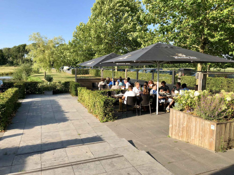 Terraza exterior con gente comiendo bajo sombrillas en Camping Betuwe, un parque vacacional en Gelderland, Países Bajos.