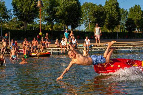 Junge springt von einem roten Schlauchboot ins Wasser, Zuschauer am Ufer bei Camping Betuwestrand, Niederlande.