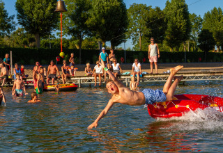 Junge springt von einem roten Schlauchboot ins Wasser, Zuschauer am Ufer bei Camping Betuwestrand, Niederlande.