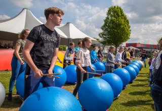 Børn deltager i en udendørs trommeworkshop med store blå træningsbolde på Camping Betuwestrand i Gelderland.