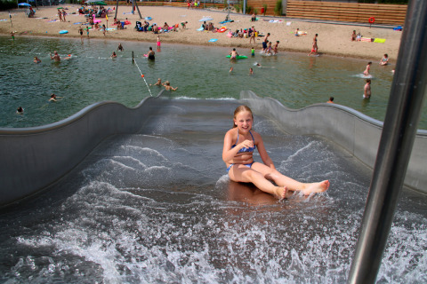 Une fille descend un toboggan aquatique à la plage d’un lac, avec des enfants jouant en arrière-plan.