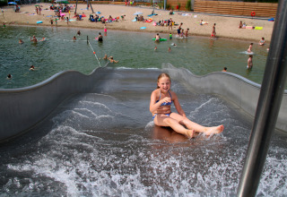 Une fille descend un toboggan aquatique à la plage d’un lac, avec des enfants jouant en arrière-plan.