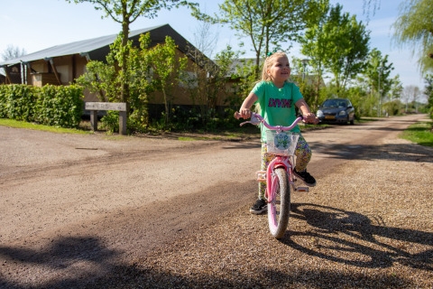 Una niña sonriente monta una bicicleta rosa en un camino de grava en el parque Camping Betuwestrand, Holanda.
