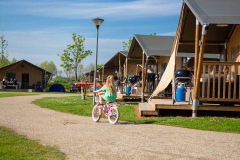 Una niña monta en bicicleta por un sendero frente a tiendas y cabañas en Camping Betuwestrand, Gelderland.