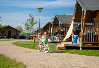 Una niña monta en bicicleta por un sendero frente a tiendas y cabañas en Camping Betuwestrand, Gelderland.