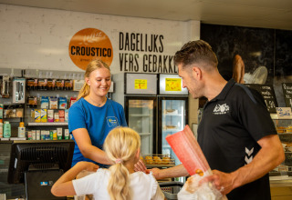 Padre e hija comprando en una tienda del camping Betuwestrand, Gelderland, Países Bajos, con cajera sonriente.