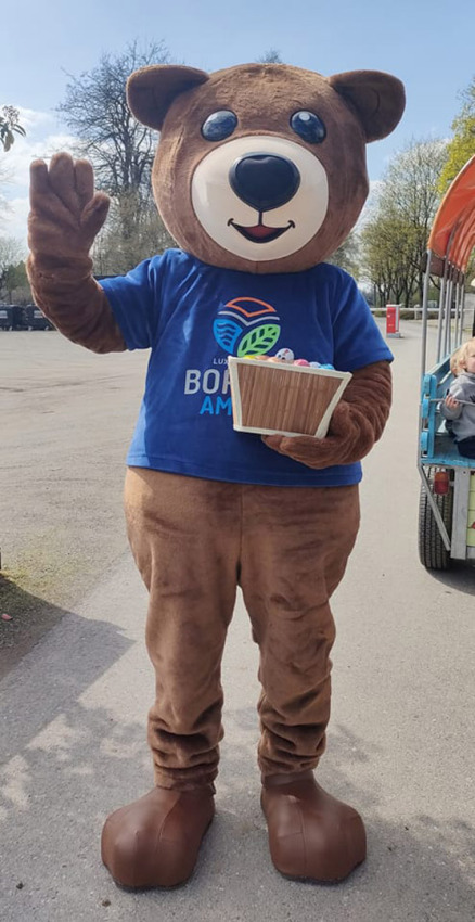 Mascota con disfraz de oso, camiseta azul y cesta saludando en Camping Borken am See, Renania del Norte-Westfalia.