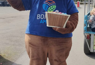 Mascotte in berenpak met blauwe T-shirt en mand zwaait op Camping Borken am See, Noordrijn-Westfalen.