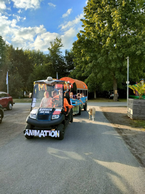 Animatiewagen met vrolijke mensen en een hond bij Camping Borken am See, omgeven door bomen.