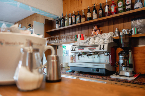 Zona de bar con cafetera, tazas, vasos y botellas en Camping Casa dei Prati, en la Toscana, Italia.