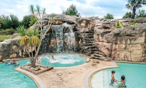Piscina con cascada, rocas y palmeras en Camping Castel L'Orangerie de Lanniron, parque vacacional en Bretaña, Francia.
