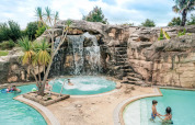 Piscina con cascata, rocce e palme al Camping Castel L'Orangerie de Lanniron, parco vacanze in Bretagna, Francia.