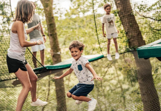 Børn og voksne leger sammen på en udendørs trampolin omgivet af træer i ferielejren L'Orangerie de Lanniron.