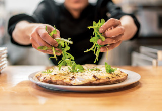 En kok pynter en friskbagt pizza med rucola på et køkken i Camping Castel L'Orangerie de Lanniron, Bretagne, Frankrig.