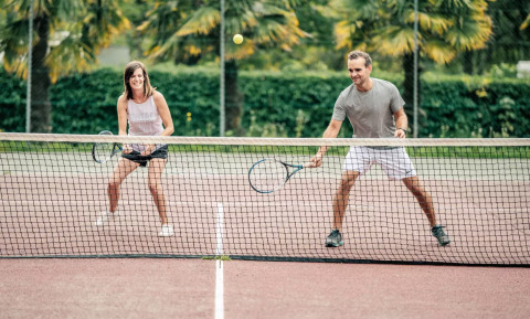 Dos adultos juegan tenis en una cancha al aire libre con palmeras en Camping Castel L'Orangerie de Lanniron, Bretaña.