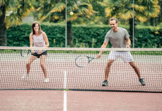 Dos adultos juegan tenis en una cancha al aire libre con palmeras en Camping Castel L'Orangerie de Lanniron, Bretaña.