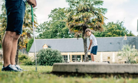 Un bambino gioca a minigolf con un adulto in un villaggio vacanze con palme in Bretagna, Francia.