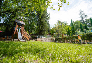 Speelplein met glijbaan en schommels op Camping Colleverde, Toscane, tussen gras en hoge bomen.