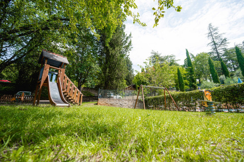 Parque infantil con columpios y tobogán en Camping Colleverde, Toscana, rodeado de césped y árboles altos.