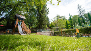 Parque infantil con columpios y tobogán en Camping Colleverde, Toscana, rodeado de césped y árboles altos.