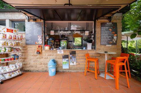 Café al aire libre en Camping Colleverde, Toscana, Italia, con estanterías de snacks, menús y sillas naranjas.