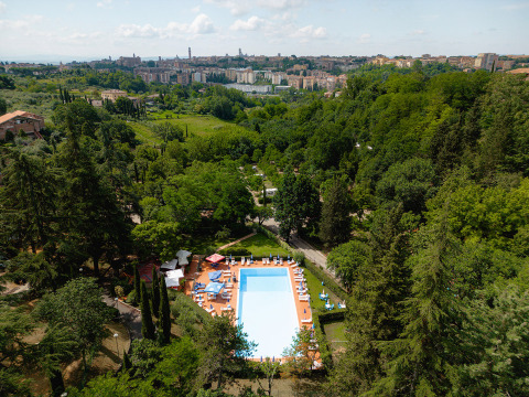 Vista aérea de una piscina en Camping Colleverde, rodeada de árboles y con Siena, Toscana, Italia, al fondo.