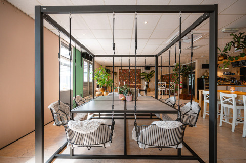 Área de comedor moderna con sillas columpio, mesa central y plantas en un parque vacacional holandés.