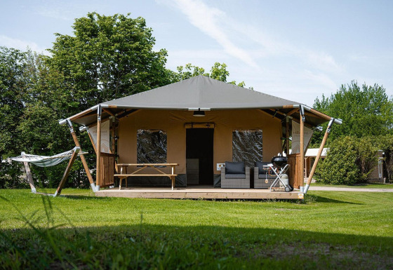 Tienda tipo safari con terraza cubierta, muebles de exterior, barbacoa y hamaca en un camping verde.