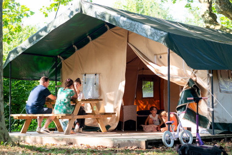 Famiglia si rilassa davanti a una tenda safari, adulti al tavolo da picnic, bambini che giocano, bici fuori.