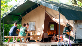 Famiglia si rilassa davanti a una tenda safari, adulti al tavolo da picnic, bambini che giocano, bici fuori.