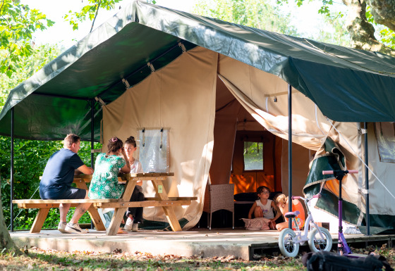 Famiglia si rilassa davanti a una tenda safari, adulti al tavolo da picnic, bambini che giocano, bici fuori.
