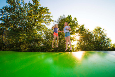 Dos niños saltan sobre una gran almohada inflable verde en Camping de Papillon, Overijssel, rodeados de árboles.